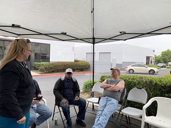 Three people chatting under a white canopy tent beside a parking lot