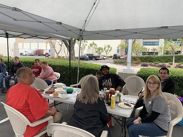 People sitting at an outdoor picnic table under a white canopy, eating and chatting near a parking lot.