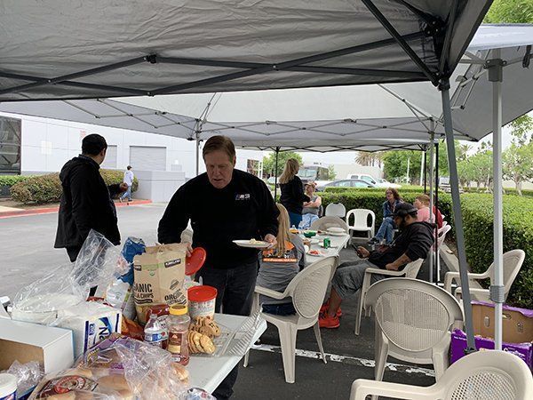 People under a canopy serving food at an outdoor gathering, with tables, chairs, and a parking lot nearby.