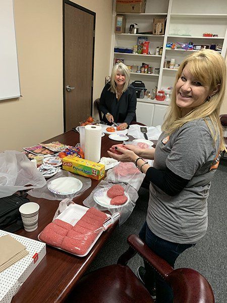 Two women preparing food at a table with plates of sliced watermelon and sandwiches in an office break room