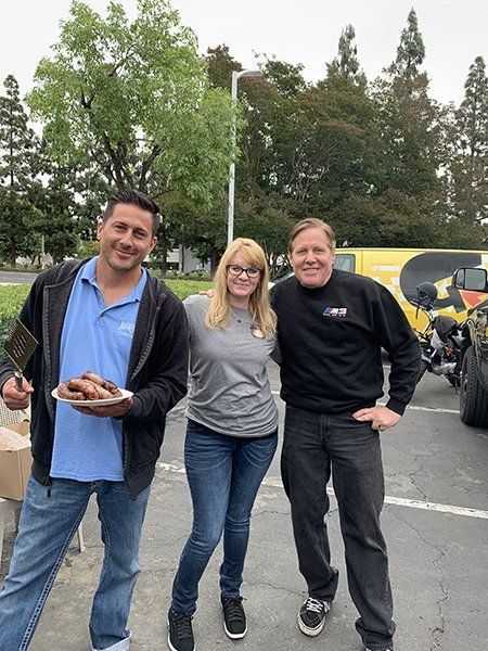 Three people smiling outdoors in a parking lot, standing near motorcycles and trees.