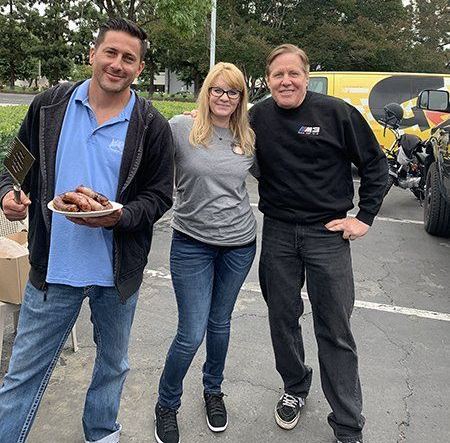 Three people posing outdoors by parked motorcycles; one holds a plate of food.