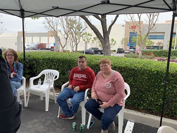 Two people seated under a canopy at an outdoor event, with hedges and chairs in the background.