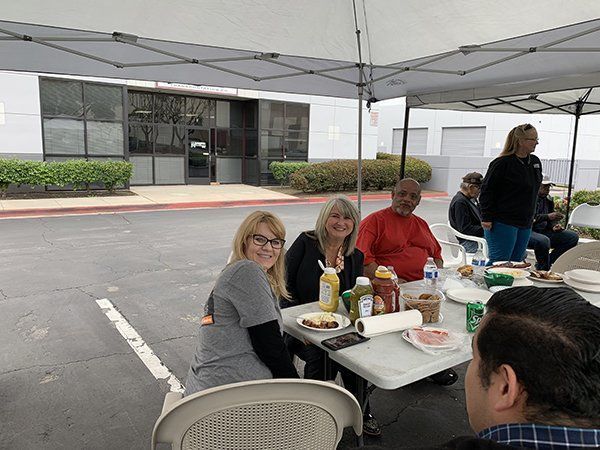 People seated at a table under a canopy, eating outdoors in a parking lot or driveway.