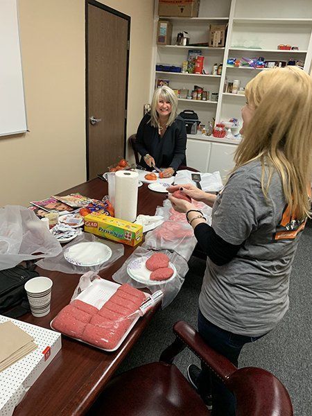 Two people stand around a table covered with food, cups, and kitchen items in a break room.
