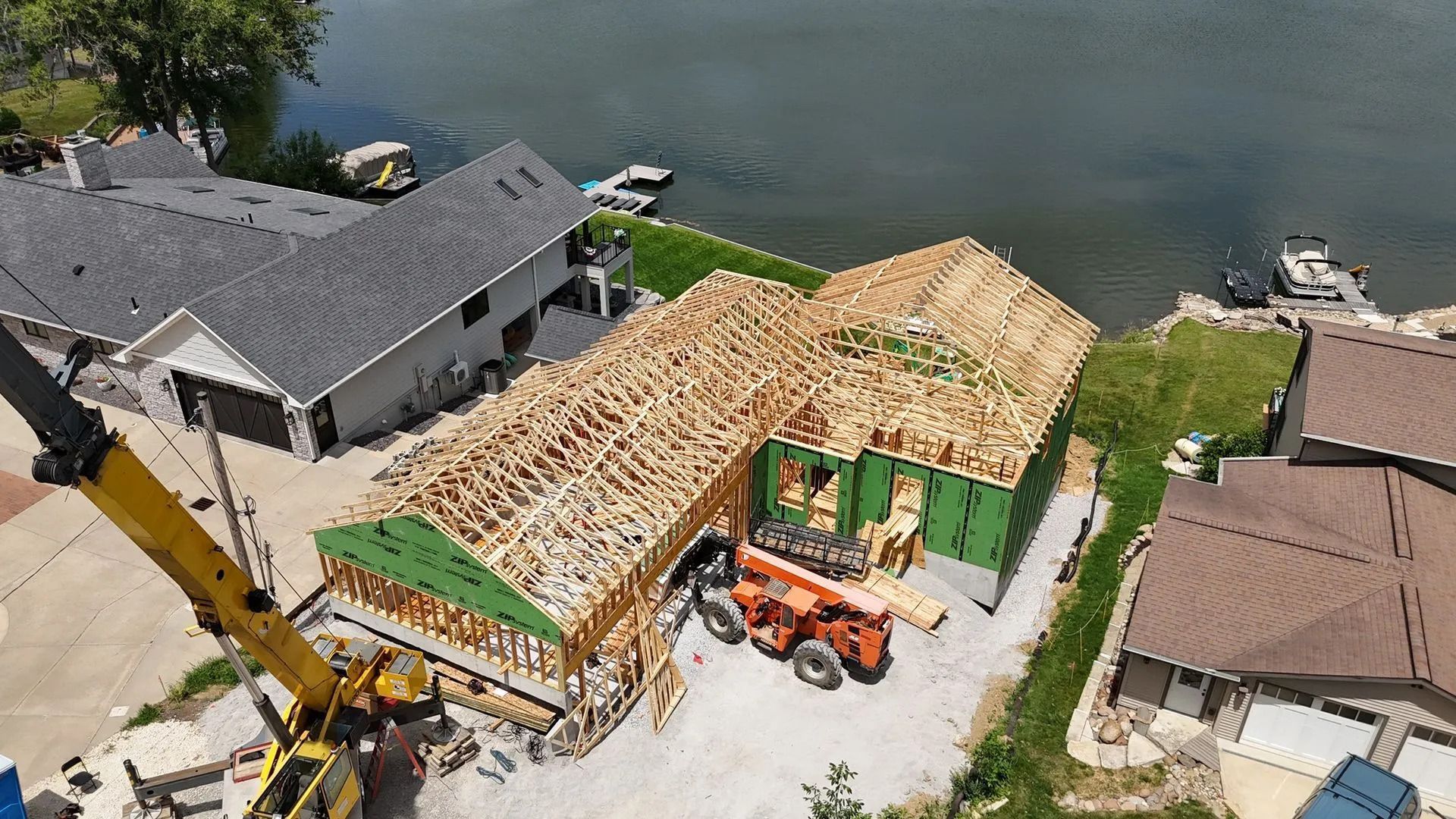 Aerial view of a house under construction by the water, with exposed roof framing, machinery, and neighboring homes.