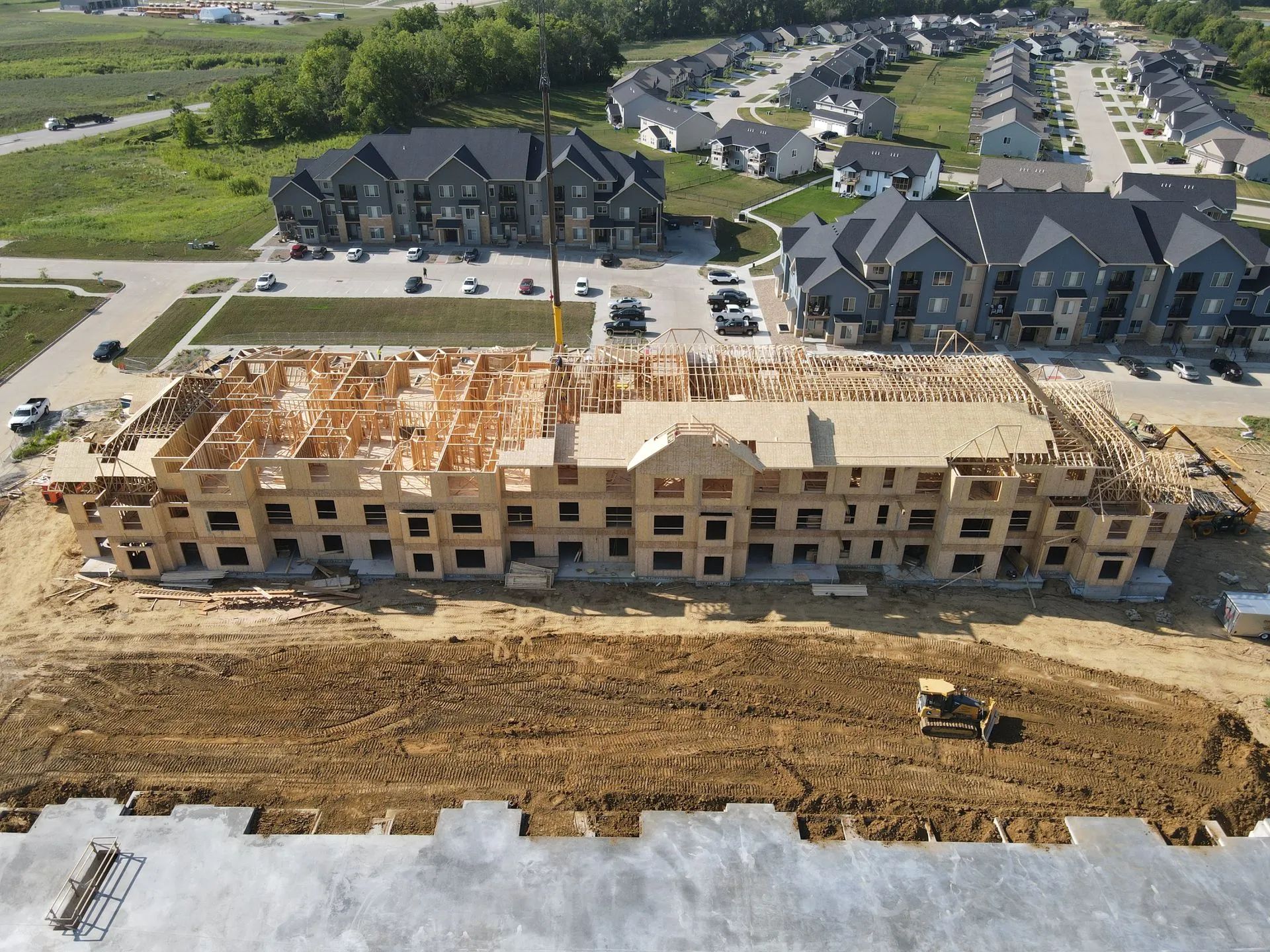 Aerial view of an apartment complex under construction beside completed homes and muddy earthworks.