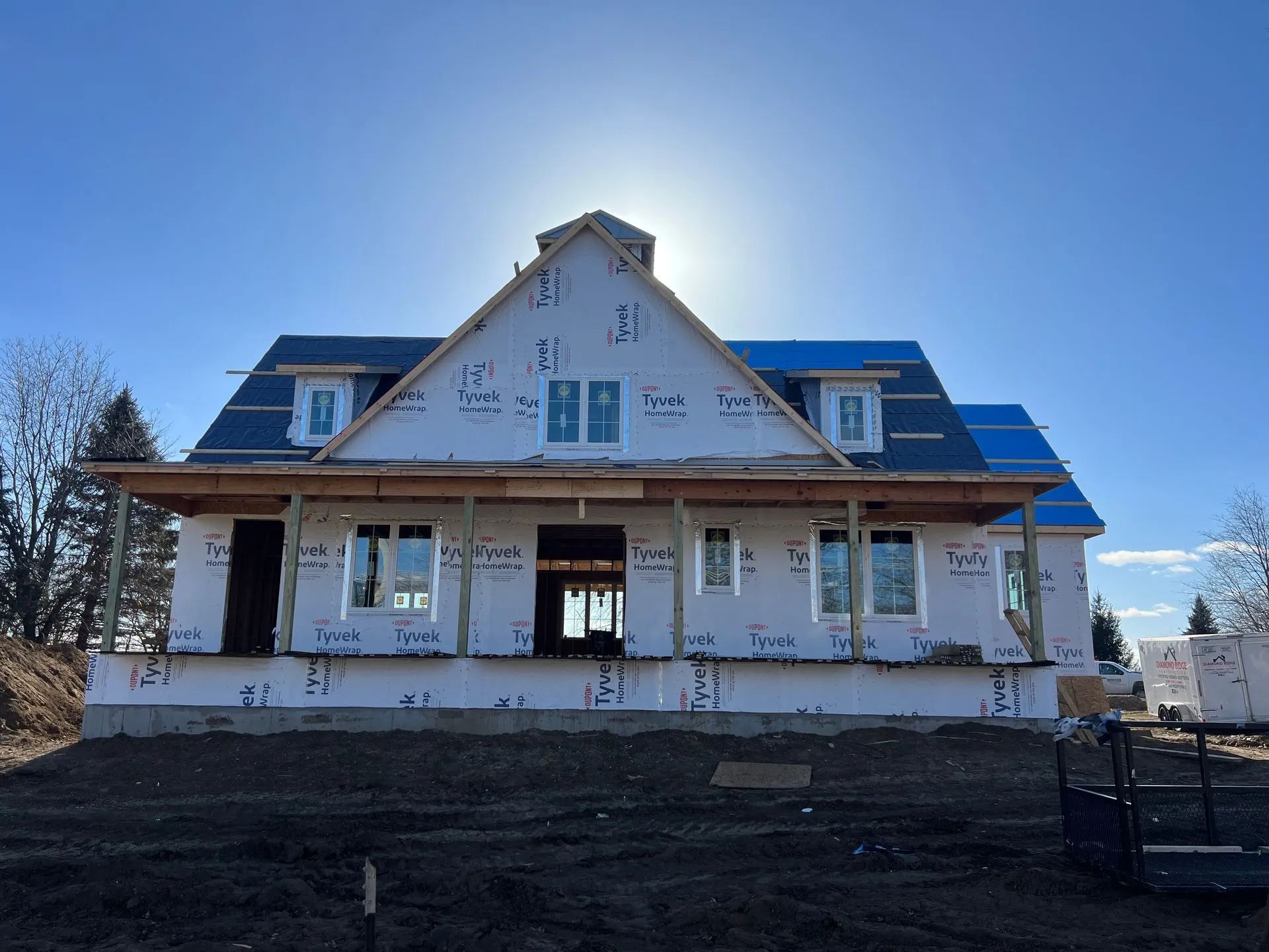 Two-story house under construction with blue roof and porch, viewed from the front in bright daylight