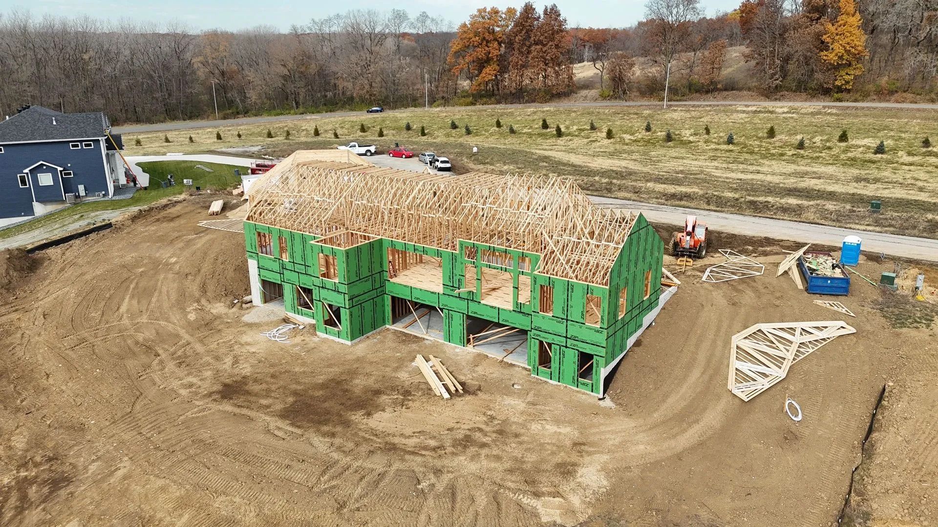 Partially built house with green framing and a straw-covered roof on a dirt lot in a rural field