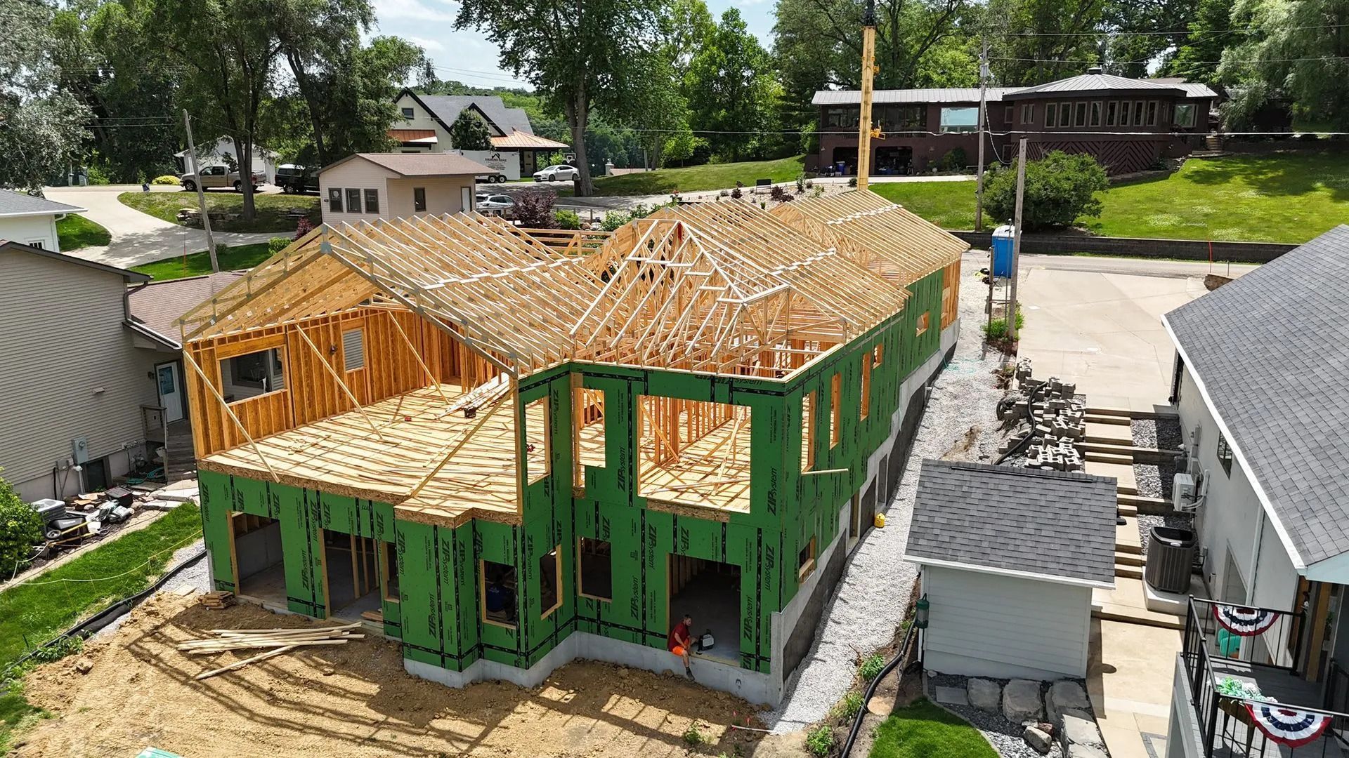Partially built house with exposed wooden framing, green exterior sheathing, and a roof under construction in a neighborhood