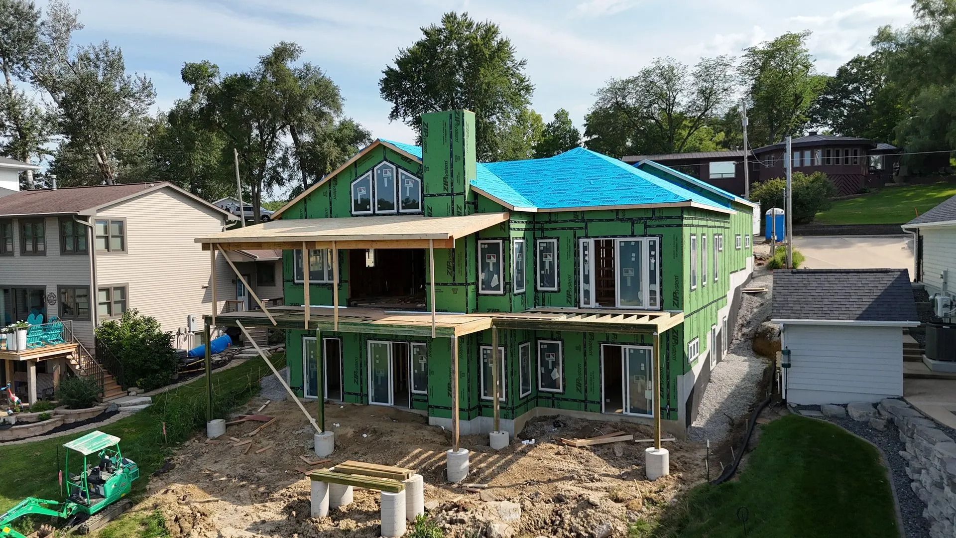 Two-story house under construction with green siding and a bright blue roof in a residential neighborhood