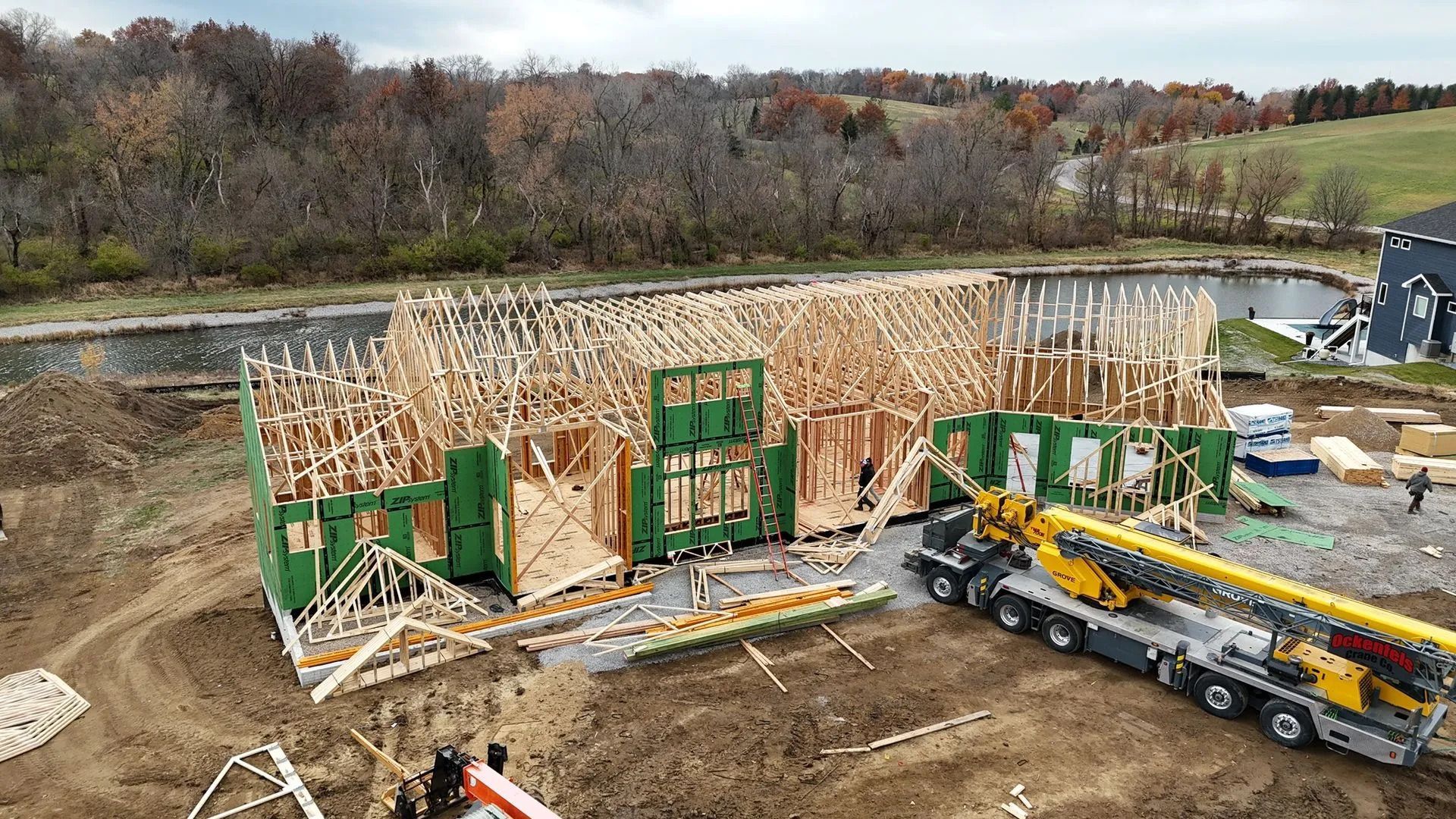 Construction site with wooden wall framing, green support panels, and a crane on muddy ground near a river.