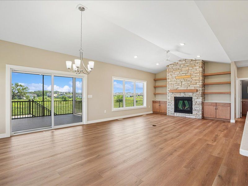 Bright empty living room with hardwood floors, stone fireplace, vaulted ceiling, and sliding glass doors to a deck