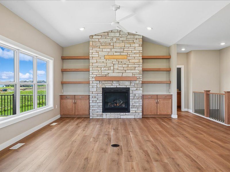 Bright living room with stone fireplace, built-in shelves, wood floors, and large windows overlooking greenery