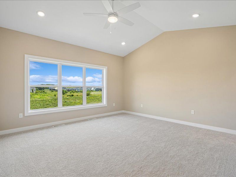 Empty beige bedroom with carpet, ceiling fan, and large window overlooking green fields