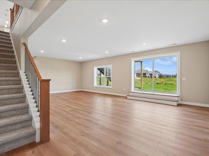Empty living room with wood floors, carpeted stairs, recessed lights, and large windows overlooking a yard