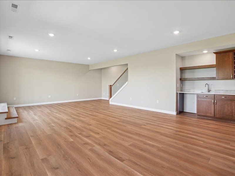 Empty open-plan living room with wood floors, white walls, recessed lights, staircase, and small kitchen area