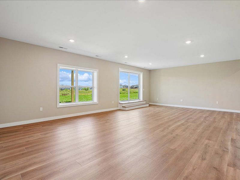 Empty living room with hardwood floors, beige walls, and large windows overlooking a green field