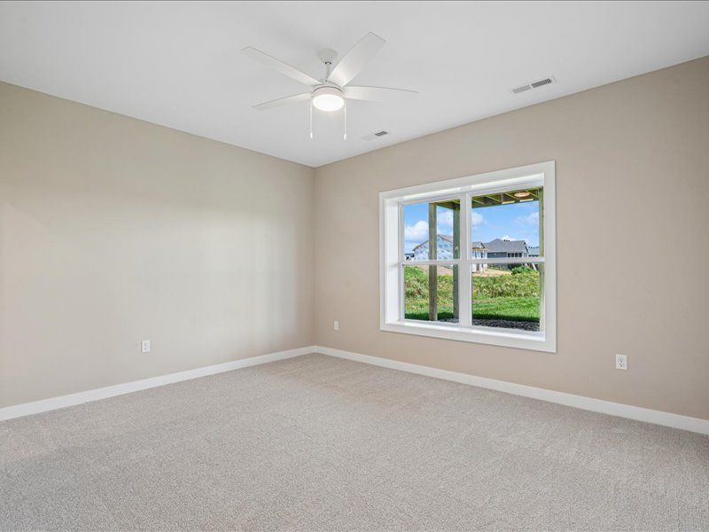Empty beige bedroom with carpet, ceiling fan, and window overlooking greenery