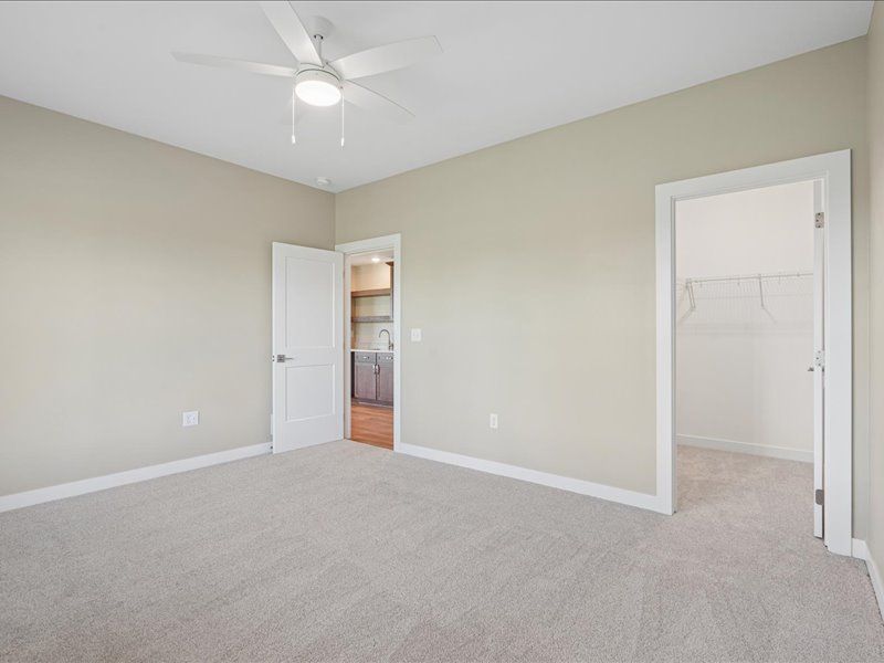 Empty beige bedroom with carpet, white ceiling fan, and two open doorways leading to a bathroom and hall.
