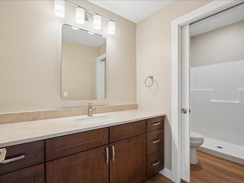 Modern bathroom vanity with mirror, beige walls, white tub, and wood cabinets beside an open doorway