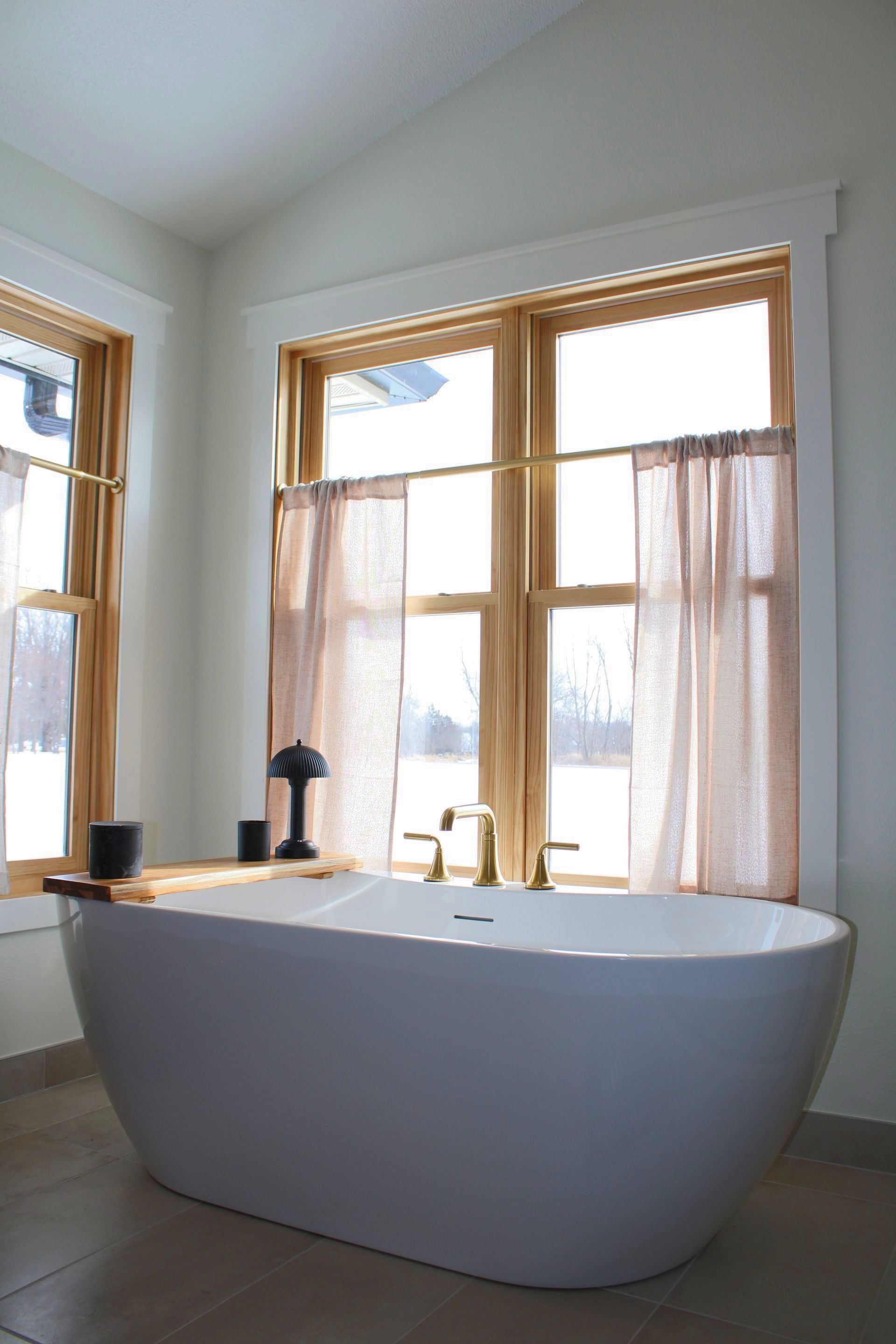 A white freestanding bathtub sits in front of windows with light curtains, brass fixtures, and a wooden tray.