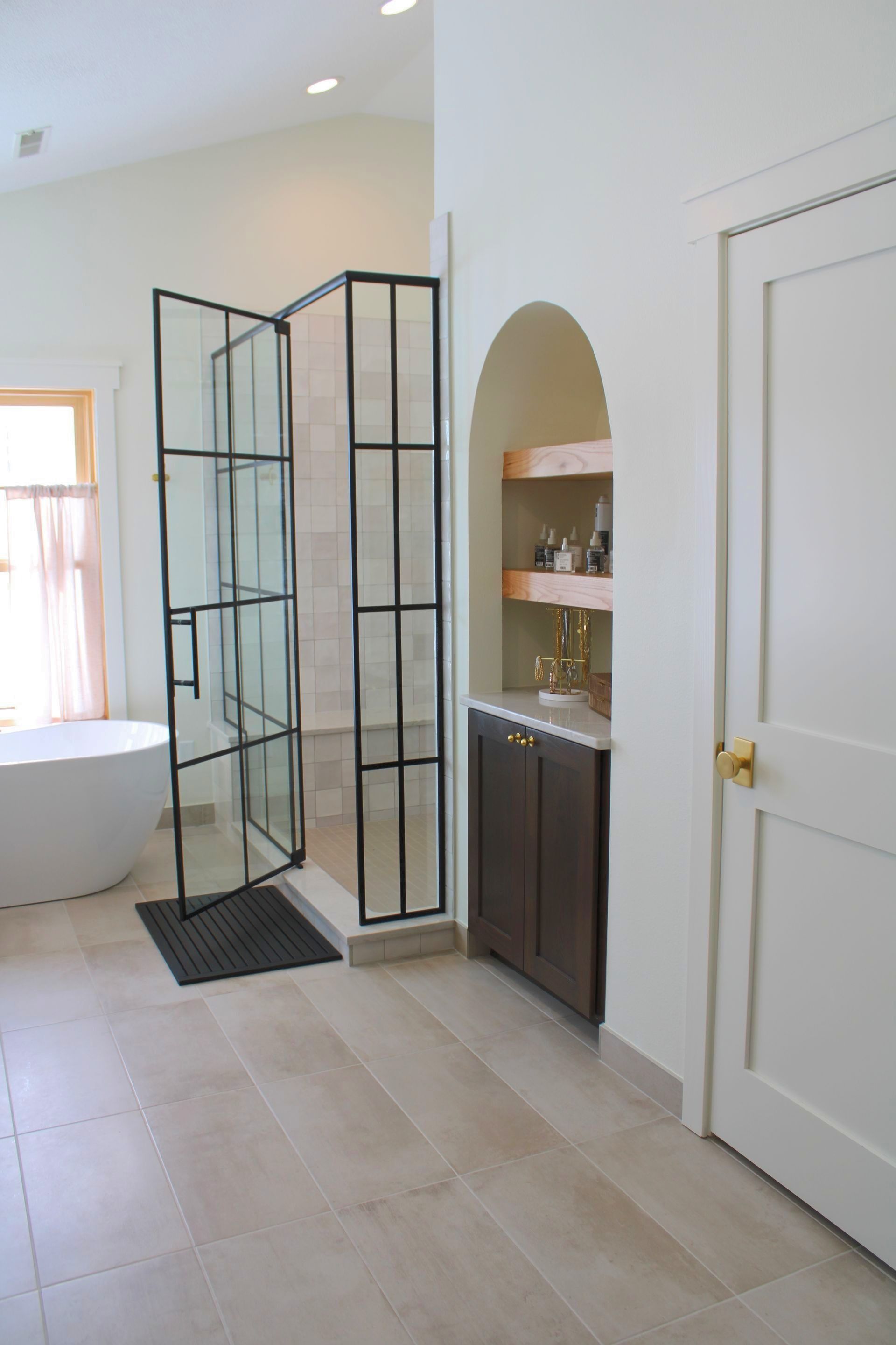A bright bathroom with a white freestanding tub, a glass-enclosed shower with black frames, and an arched storage nook.