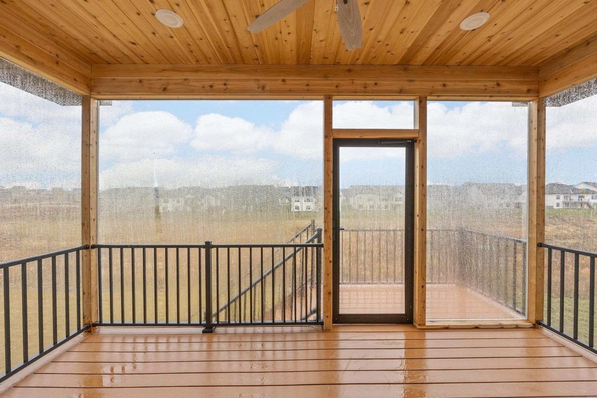 A covered wooden porch with a black railing, ceiling fan, and a screened door leading to an outdoor deck.