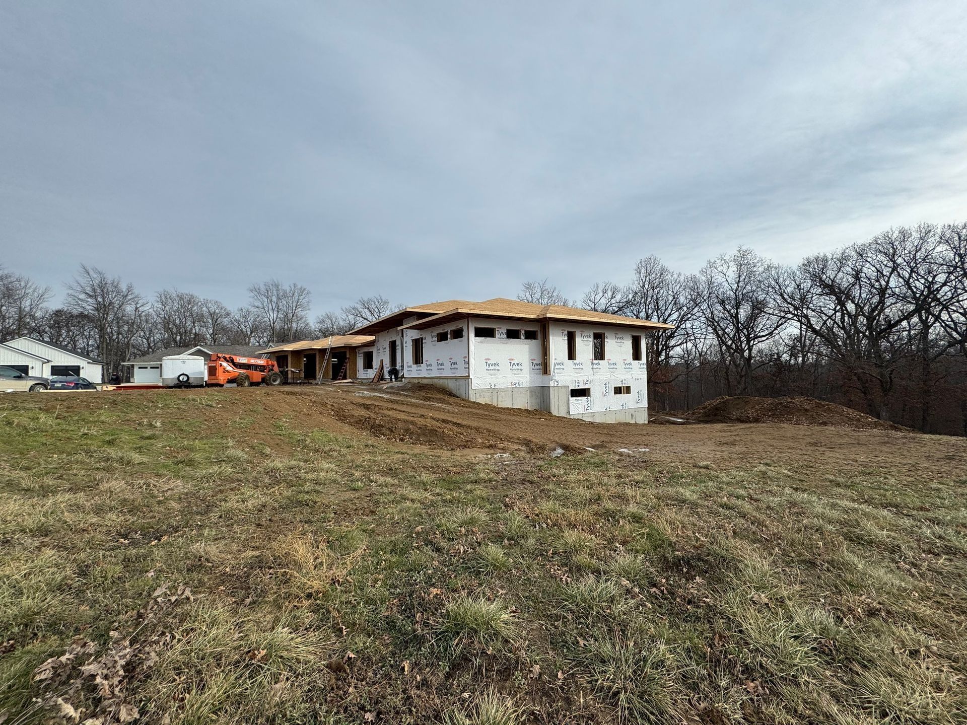 A house under construction with white exterior panels, framed by trees on a cloudy day over a grassy, dirt-covered hill.