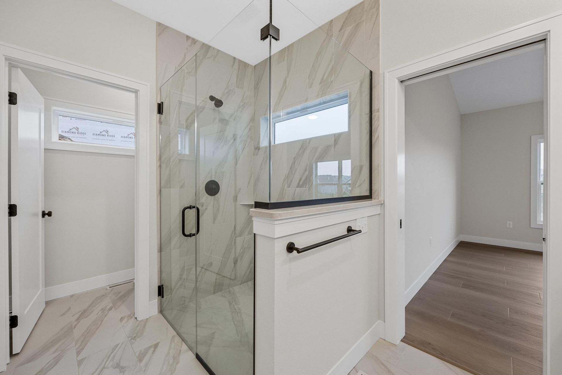 A walk-in shower with marble-look tile, glass door, and black hardware between two doorways in a bright room.