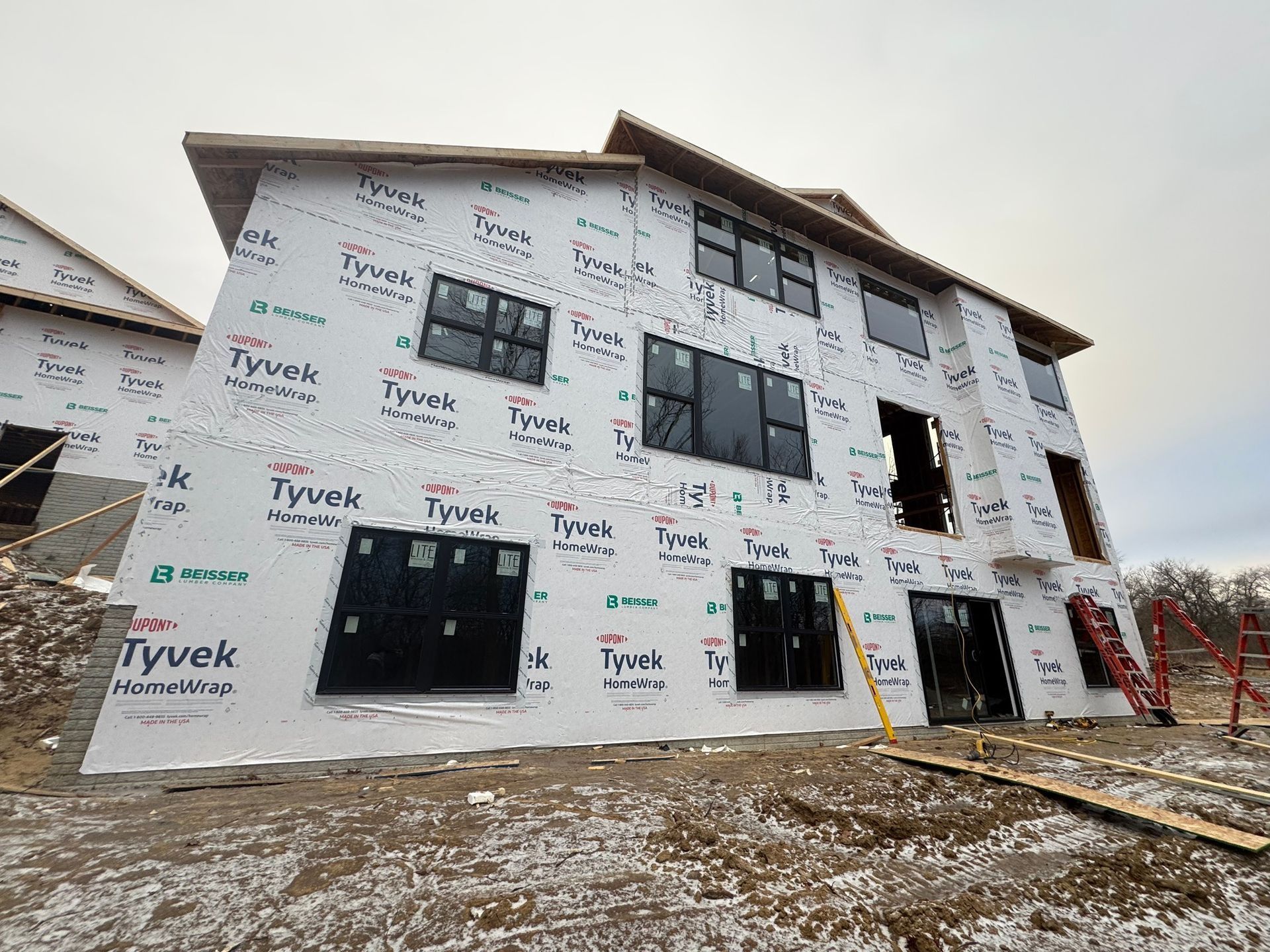 A multi-story house under construction, wrapped in white Tyvek weather-resistant barrier, with dark-framed windows.