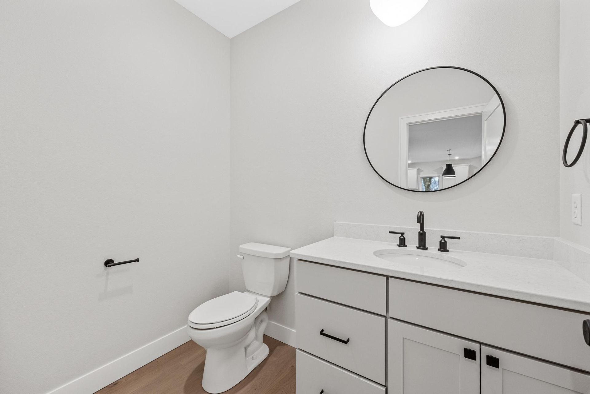A modern bathroom with light gray walls, a white vanity with black hardware, a round mirror, and a white toilet.