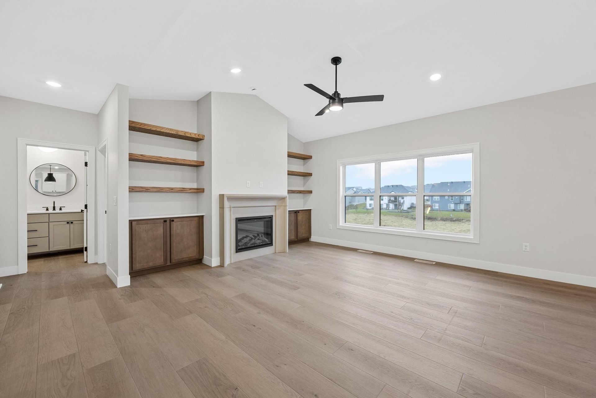 A modern living room with a fireplace, open wood shelves, light wood floors, a ceiling fan, and a view of a neighborhood.