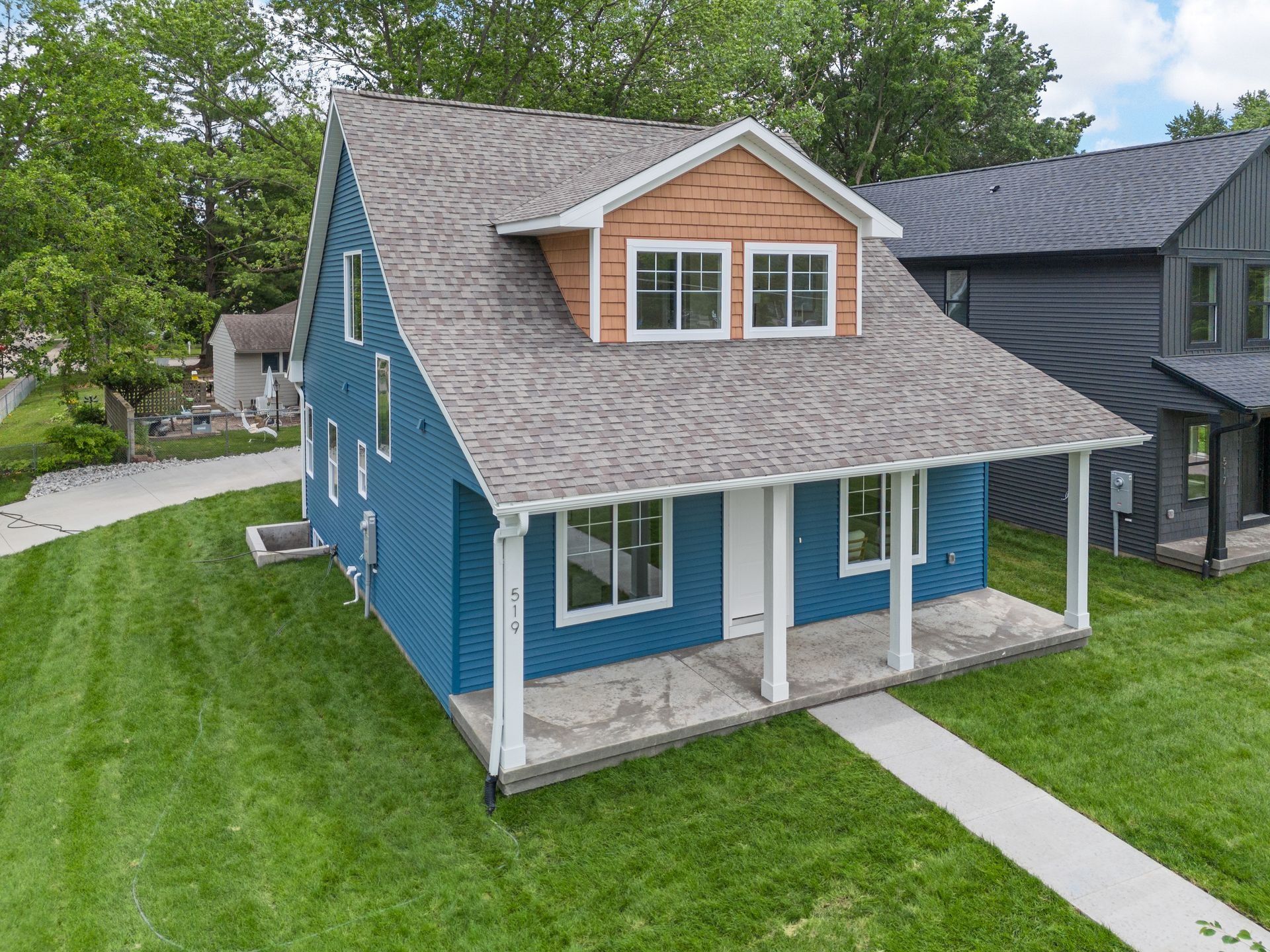 Blue two-story house with a wooden dormer, gray shingled roof, and a front porch, viewed from a high angle on a lawn.