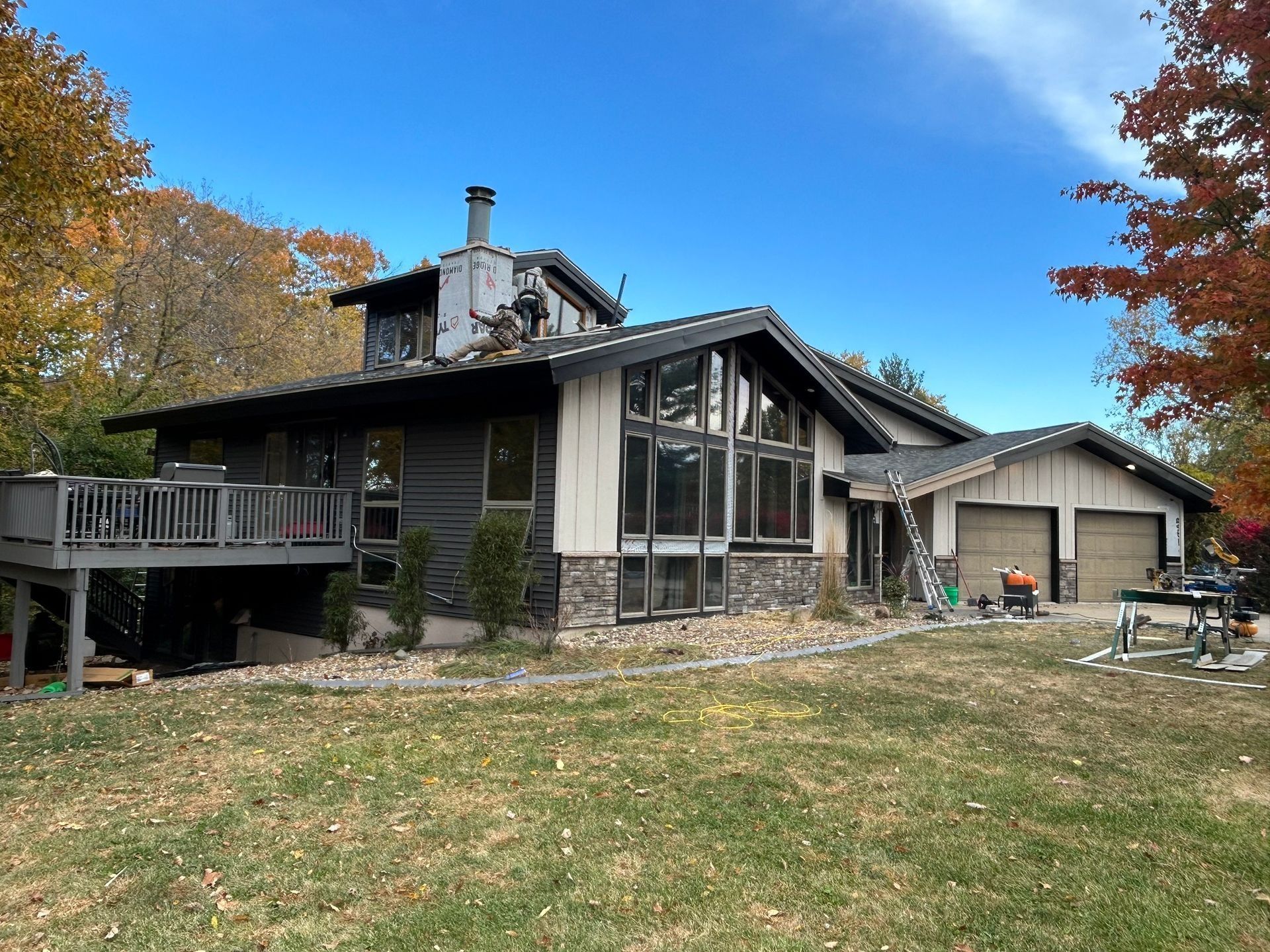 A modern, two-story house with dark siding and stone accents, a large deck, and a multi-car garage on a lawn with trees.