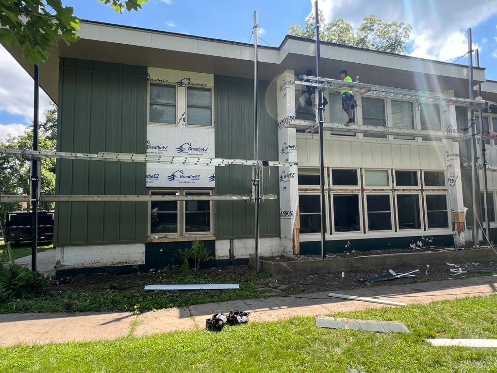 Construction workers on scaffolding repair the exterior of a two-story building with green siding and white trim.