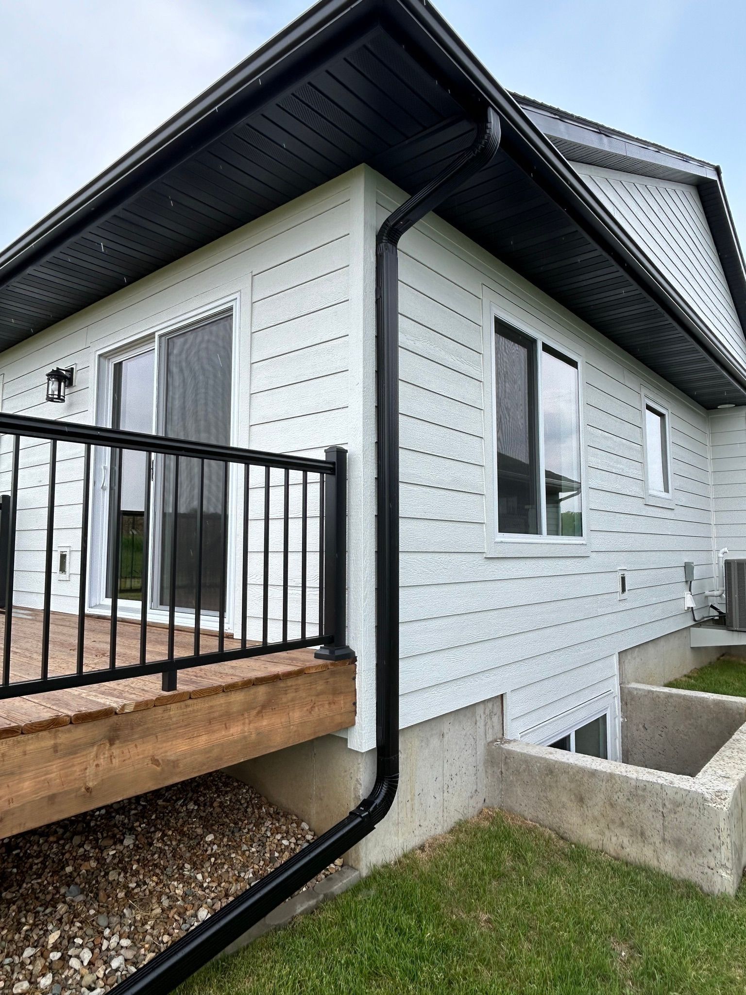 White house exterior with a deck, a black gutter downspout, windows, and a concrete window well on a lawn.
