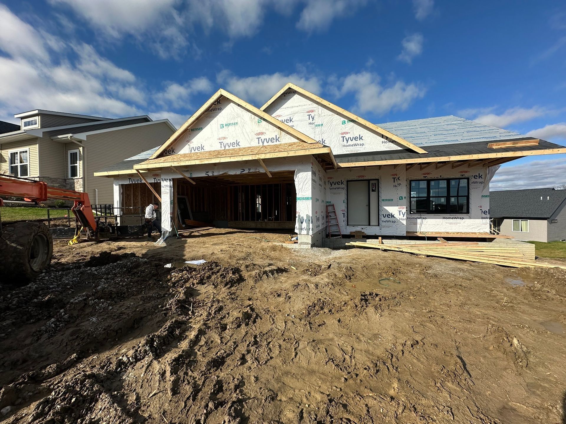 A house under construction with white protective wrap on its walls and a muddy yard under a bright, partly cloudy sky.