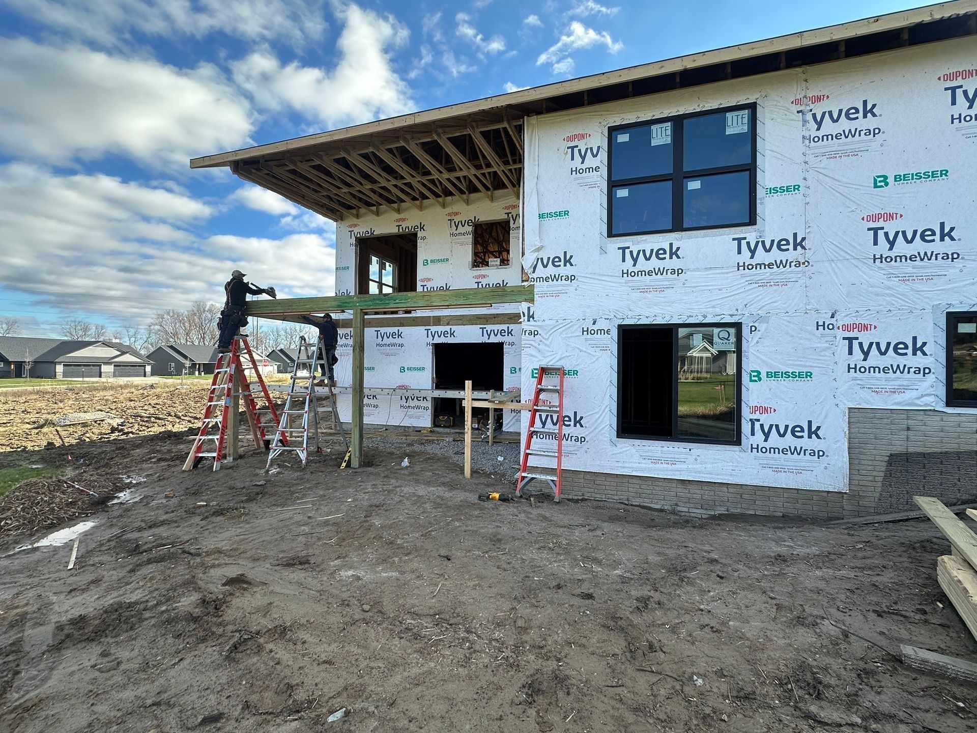 Two construction workers install a support beam on the exterior of a house under construction with Tyvek house wrap.