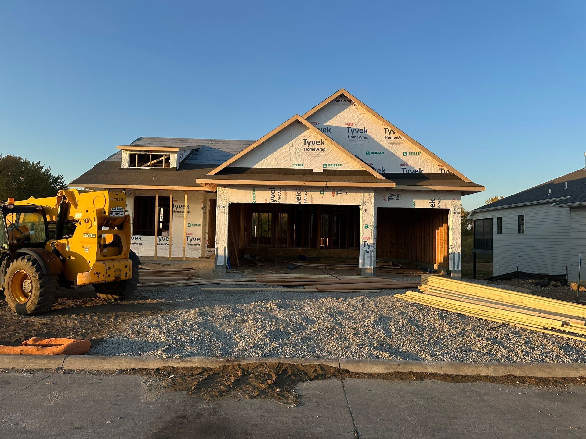 A yellow construction lift sits parked in front of a house under construction with white sheathing and a dark roof.