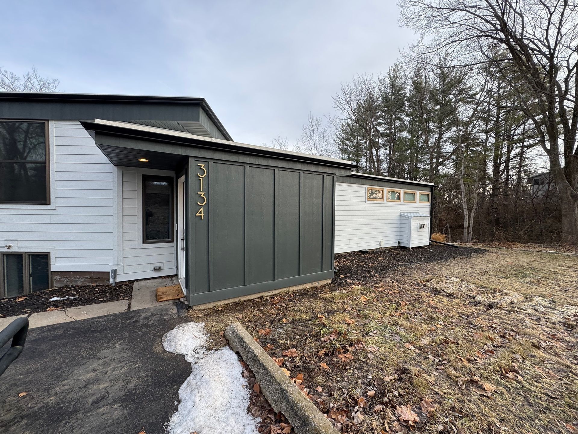 A house exterior features a white-sided building with a dark grey, modern entryway and a partially snow-covered driveway.