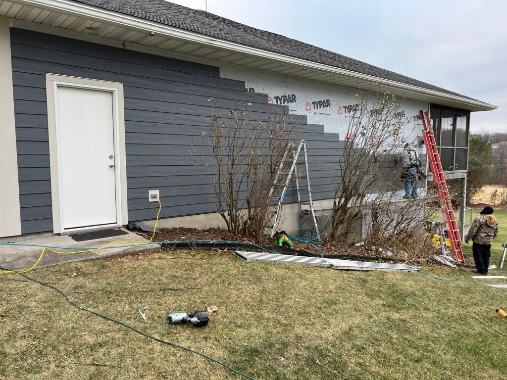 Workers install dark grey siding on a house wall, with ladders, tools, and a person standing in the yard nearby.