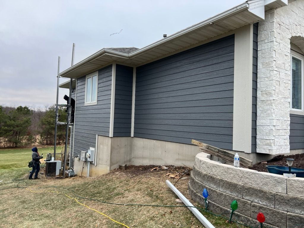 Workers use scaffolding to install dark gray horizontal siding on the side of a house with a stone-accented facade.