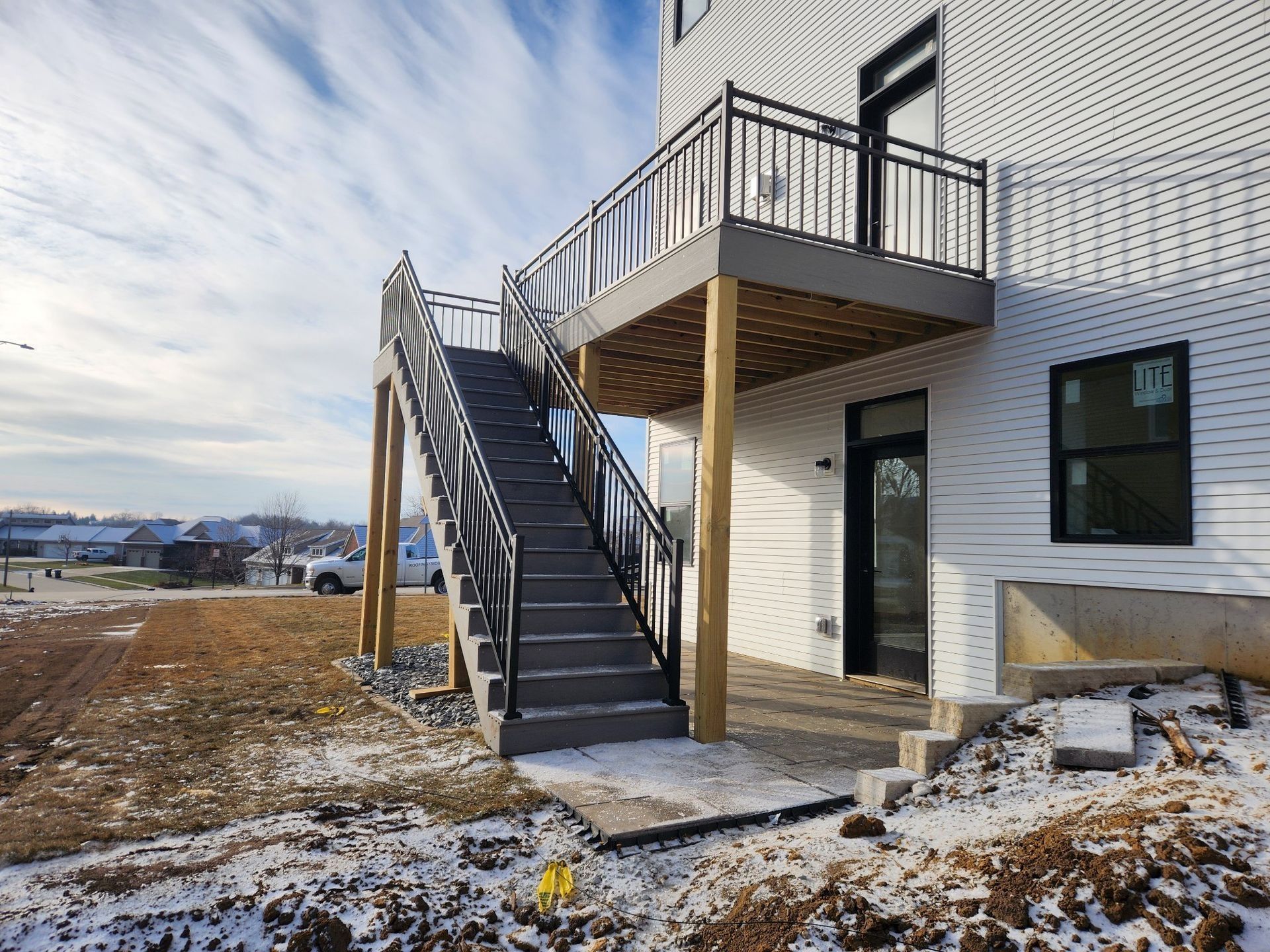 Exterior stairs leading from a ground-level concrete patio to a second-story wooden deck on a white-sided building.