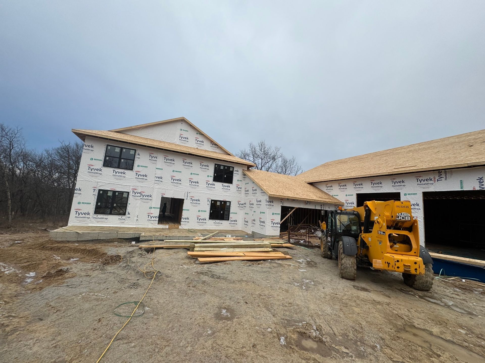 A half-built two-story house with white exterior sheathing and a yellow telehandler parked in the foreground.