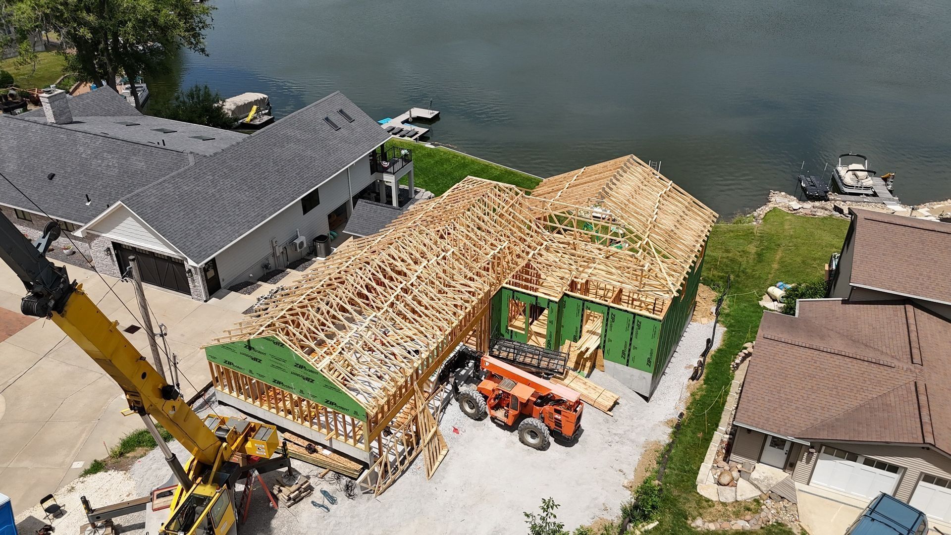 Aerial view of a residential construction site with a wooden roof frame, a yellow crane, and an orange boom lift.