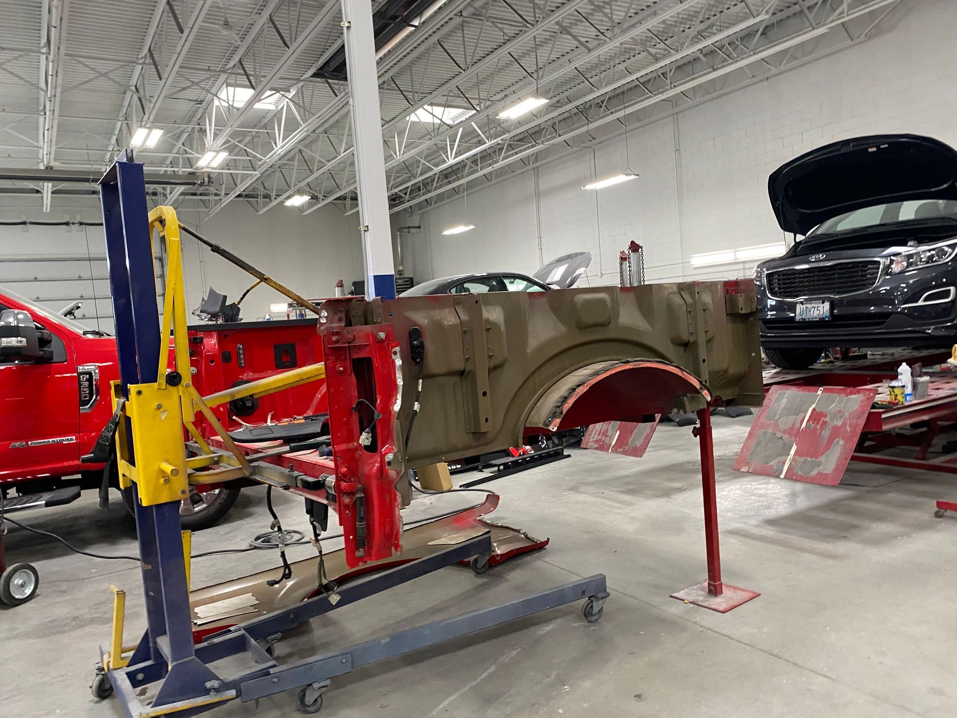 A red and metal vehicle side panel mounted on a repair stand inside a busy automotive body shop.