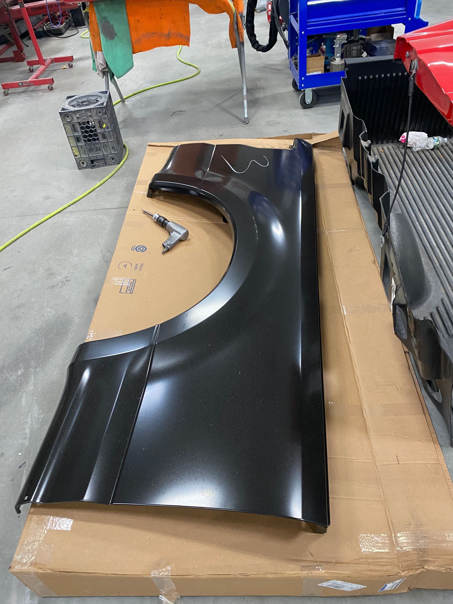 A black replacement car fender sits on cardboard in an auto body shop, with a small drill resting nearby.