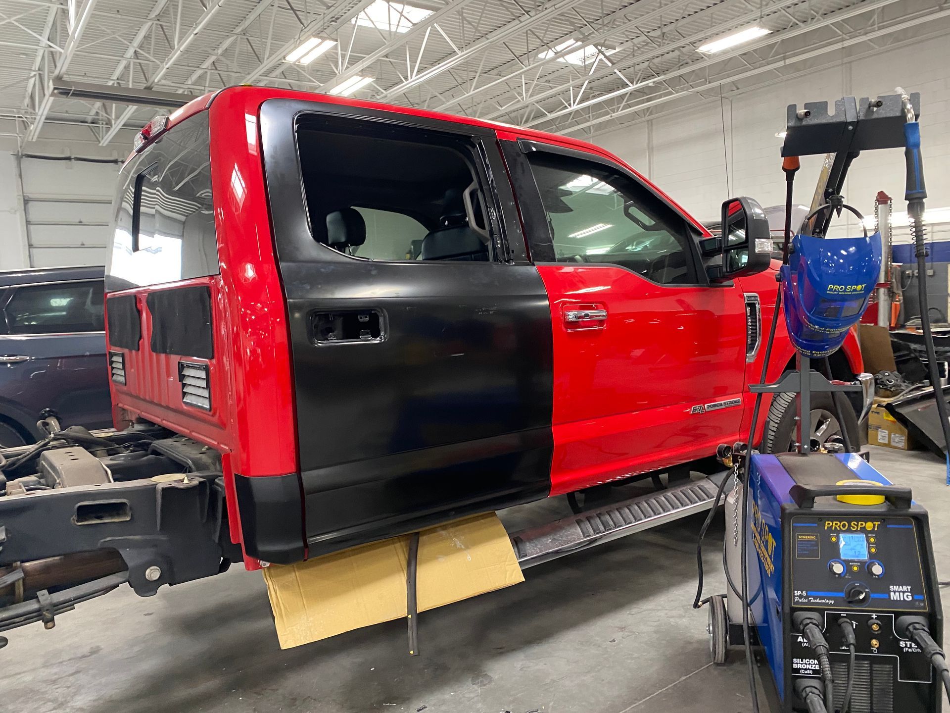 A red truck cab with a black replacement door in a repair shop with welding equipment nearby.