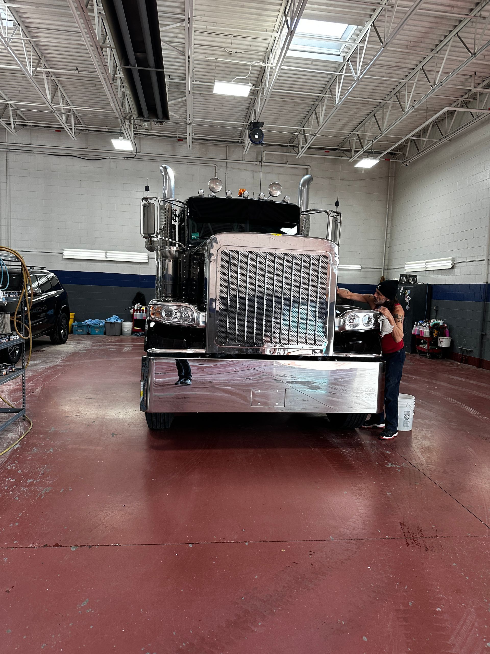 A person polishing the chrome grill and bumper of a large semi-truck inside a maintenance garage.
